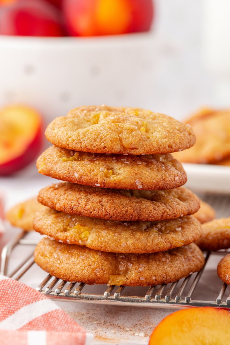 a stack of five peach cookies on a wire rack with fresh peaches in the background