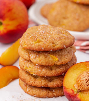 a stack of five peach cookies on a white surface with more cookies and fresh peaches in the background