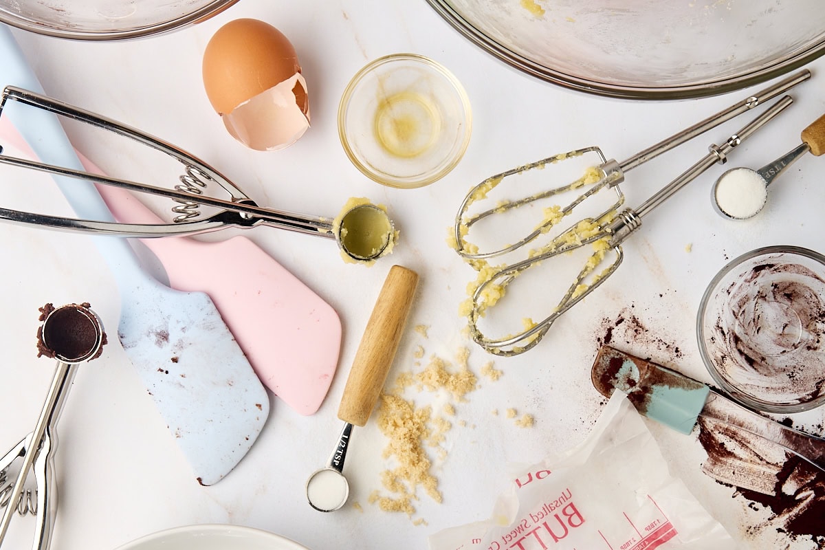 overhead view of multiple baking tools and ingredients, including beaters, a broken egg, measuring spoons, and cookie scoops