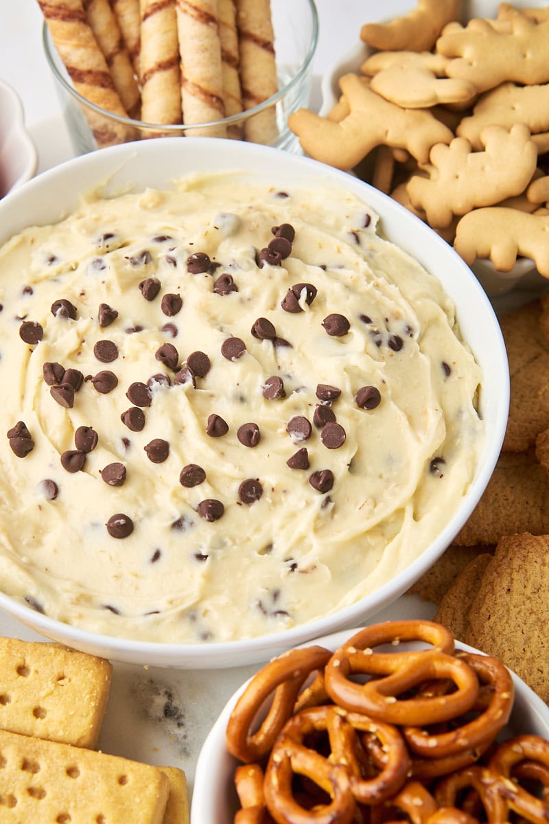 a bowl of cookie dough dip in a white bowl surrounded by pretzels, animal crackers, and more dippers