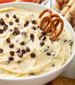 cookie dough dip in a white bowl with two pretzels sticking out; apple slices and cookies for dipping partially visible in the background