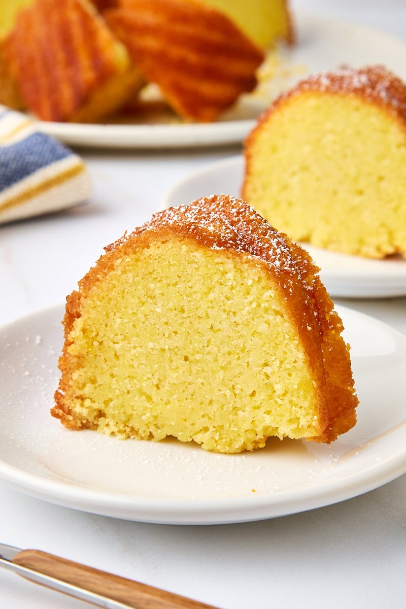 a slice of cornmeal pound cake topped with powdered sugar on a white plate with more cake in the background