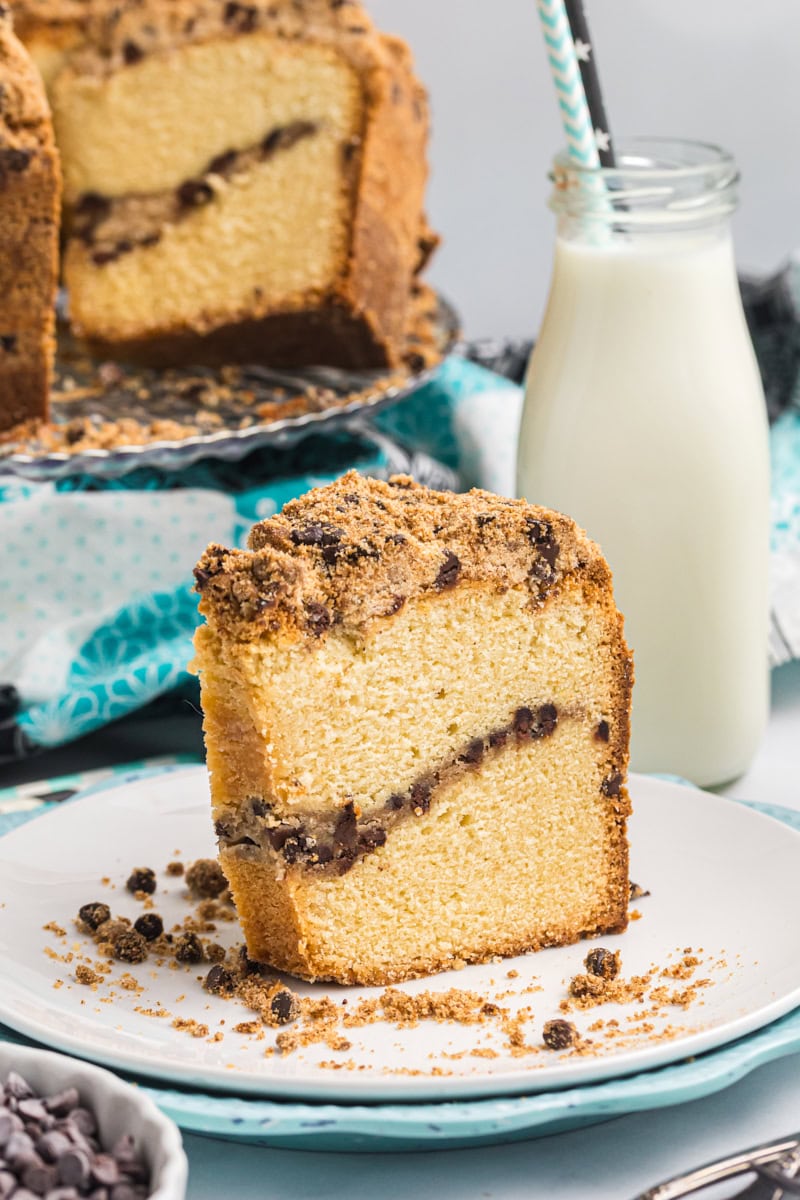 a slice of chocolate chip crumb pound cake on a white plate with remaining cake and a bottle of milk in the background