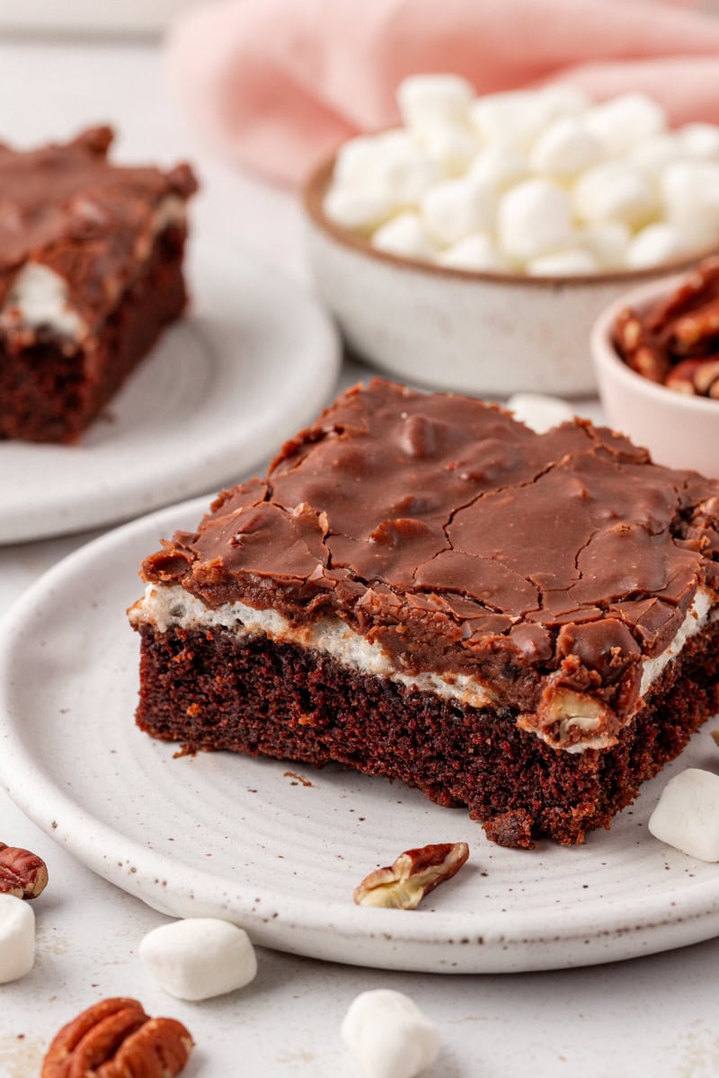 a slice of Mississippi mud cake on a white plate with more cake, pecans, and marshmallows in the background