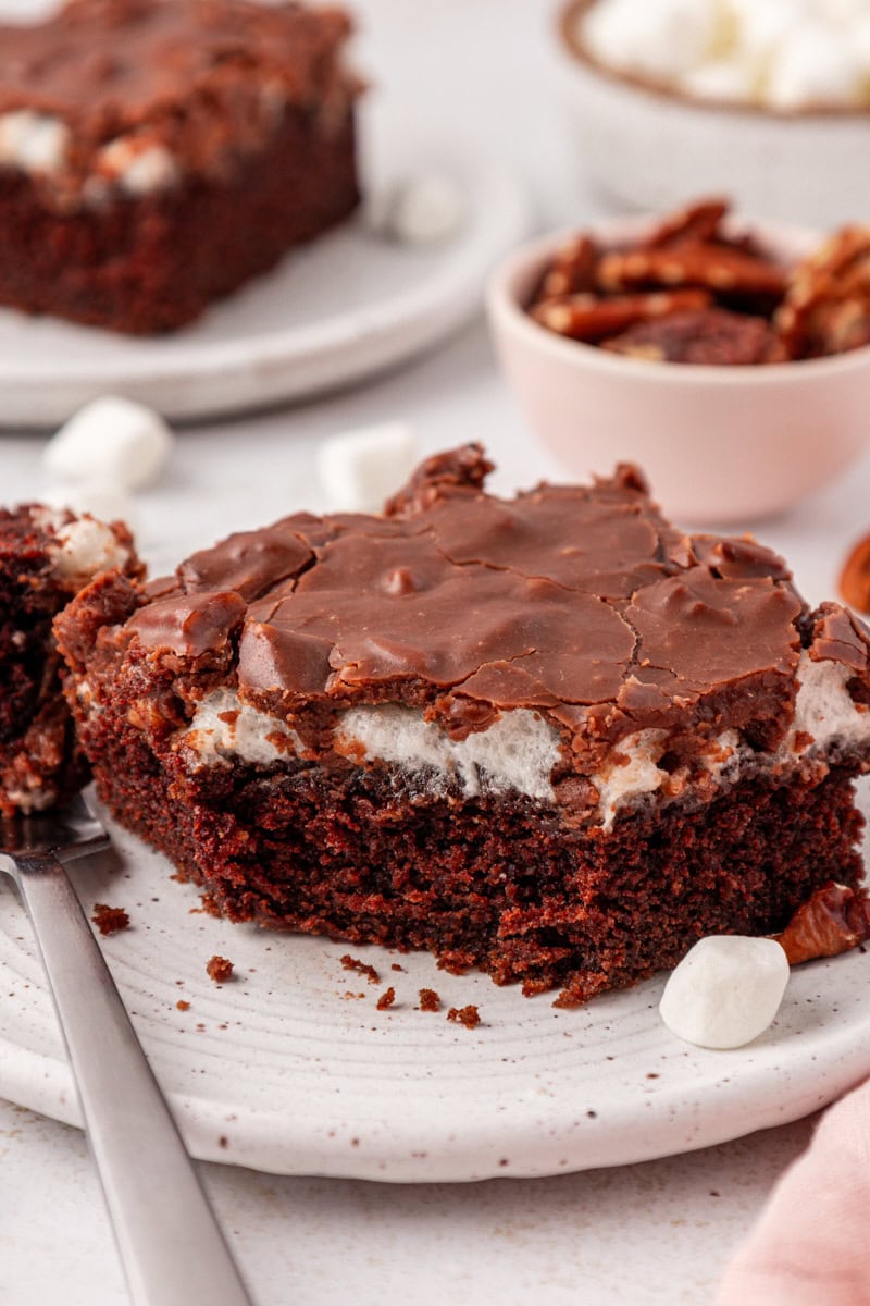 a slice of Mississippi mud cake on a white plate with a bite on a fork; more cake, pecans, and marshmallows in the background