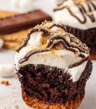 a s'mores cupcake with a bite missing with another cupcake, graham crackers, chocolate, and marshmallows partially visible in the background