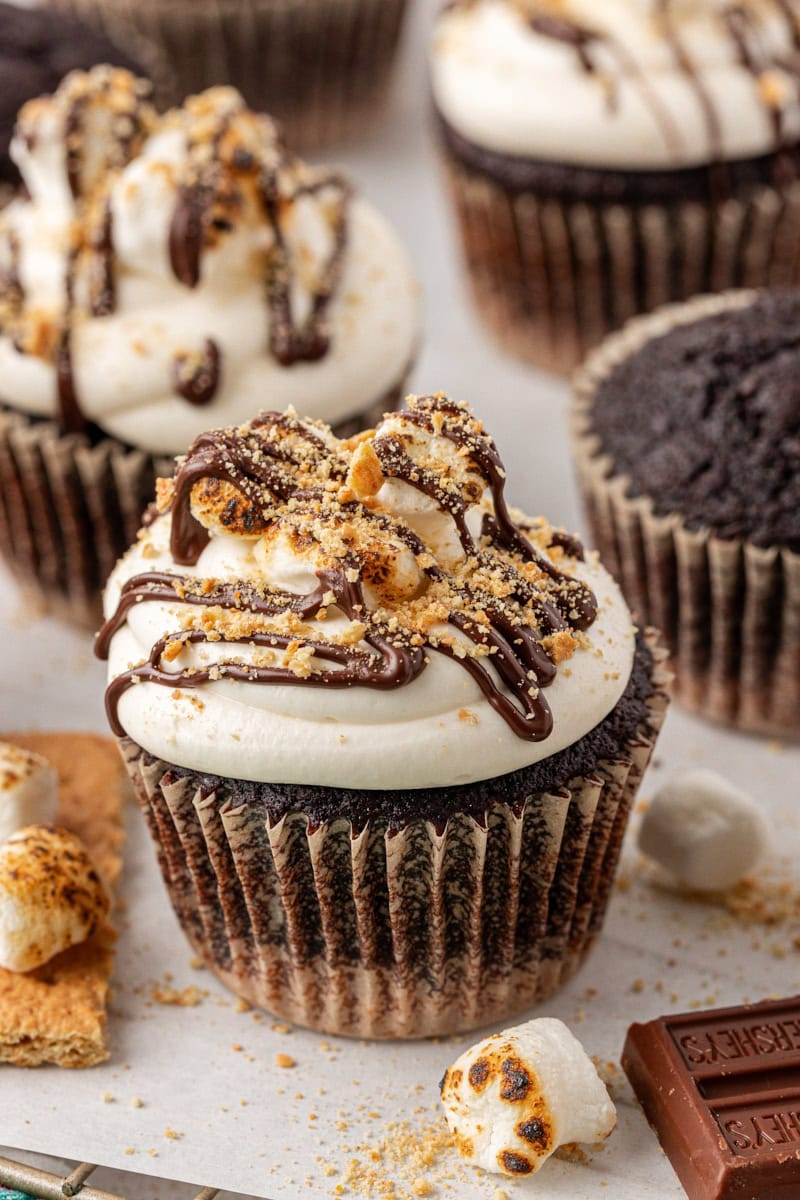 a s'mores cupcake topped with a drizzle of chocolate, toasted marshmallows, and graham cracker crumbs; more cupcakes in the background