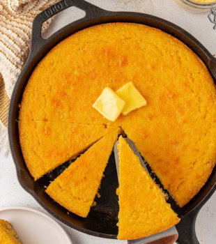 a slice of sour cream corn bread being removed from the remaining corn bread in a cast iron skillet