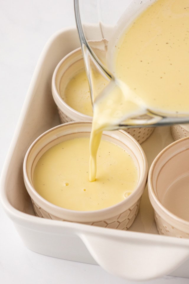 vanilla bean pots de crème mixture being poured into ramekins sitting inside a white baking dish