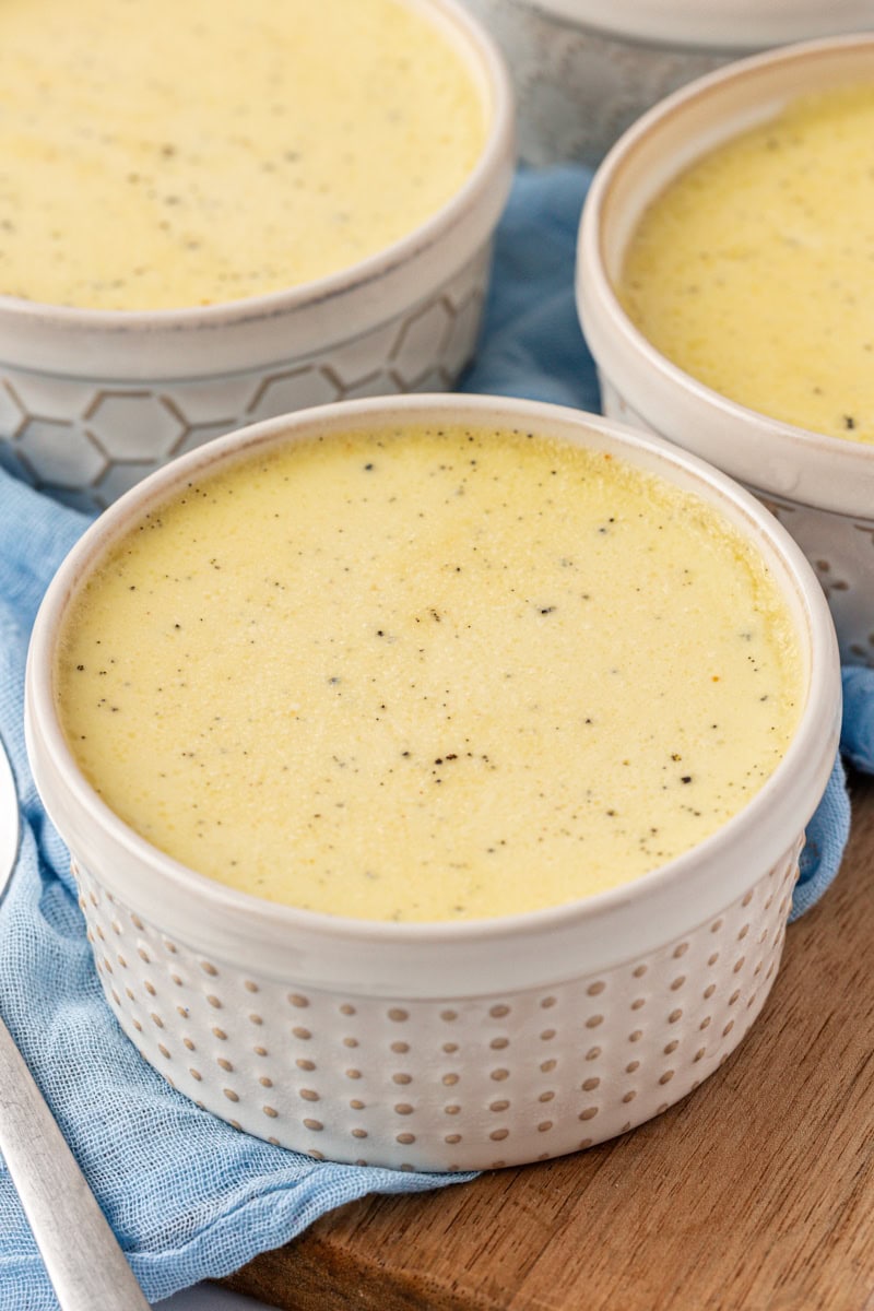 a vanilla bean pots de crème in a ramekin sitting on a wooden surface with two more servings in the background