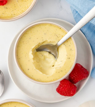 overhead view of a vanilla bean pots de crème with a spoon resting inside, set on a white plate with two fresh raspberries