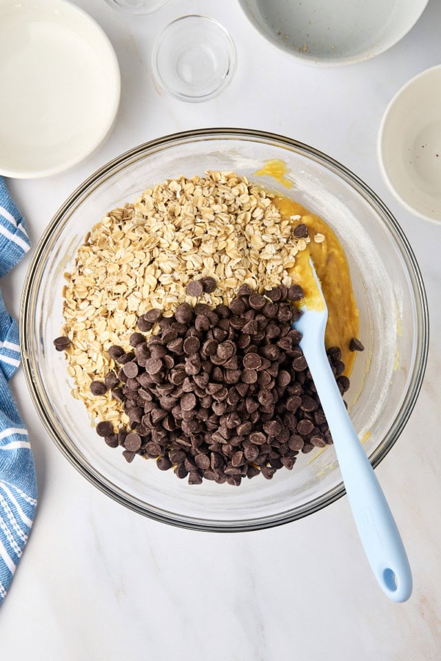 overhead view of oats and chocolate chips added to cookie batter in a glass mixing bowl