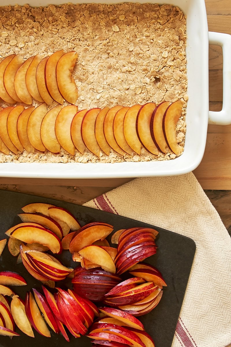 overhead view of partially assembled plum crumble bars in a white baking dish and sliced plums on a cutting board