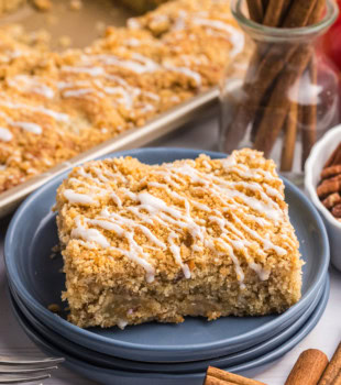 a slice of apple crumb sheet cake on a blue plate with the remaining cake in a half sheet pan in the background