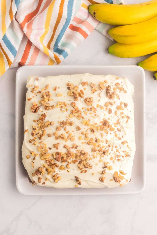 overhead view of banana cake with cream cheese frosting topped with toasted chopped nuts and served on a square white plate