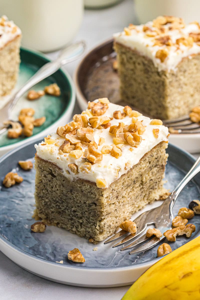 a slice of banana cake with cream cheese frosting on a blue plate with more servings in the background