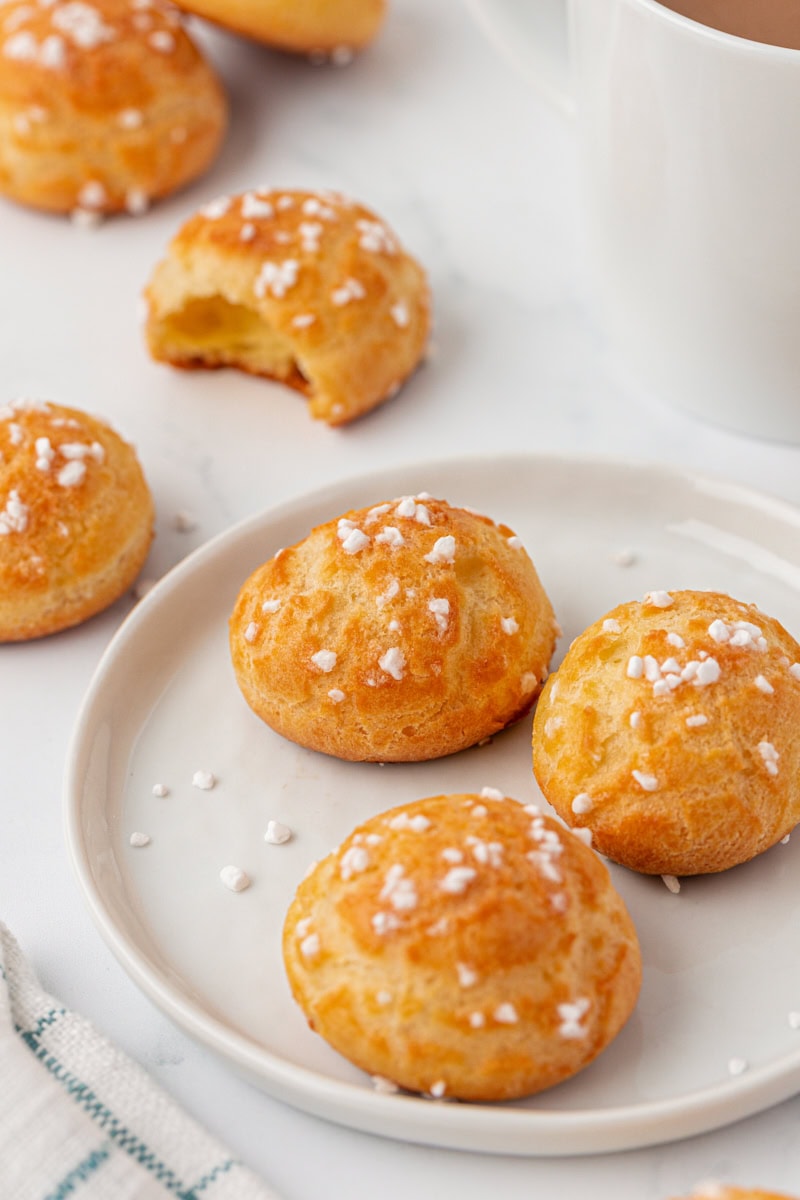 three chouquettes on a white plate with more pastries and a mug of coffee in the background