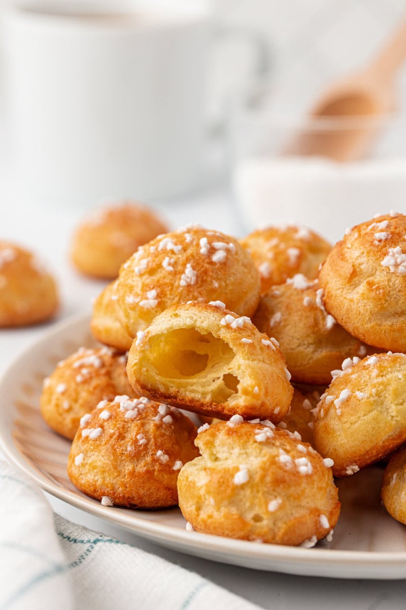 A broken-open chouquette showing the light, airy center with a crisp golden shell, sitting atop more chouquettes arranged on a white plate