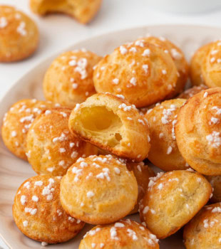 chouquettes stacked on a white plate with a bite-sized piece on top showing the inside