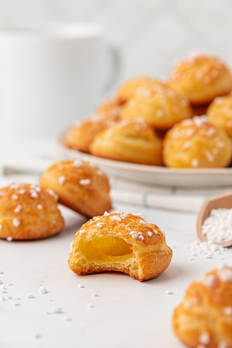 a broken-open chouquette showing the light, airy center, surrounded by more chouquettes on a white surface and on a white plate in the background