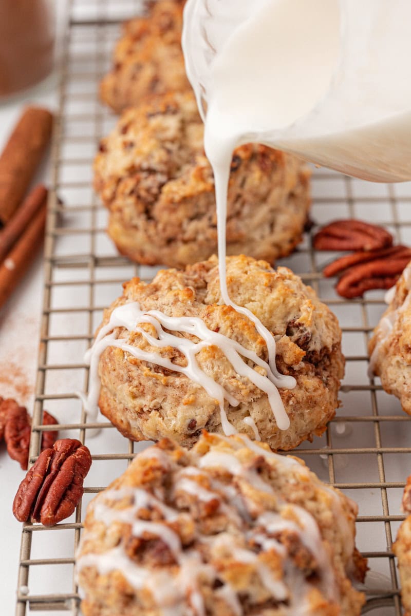 glaze being poured over a cinnamon bun scone on a wire rack; more scones and pecan halves surrounding