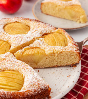 partially sliced German apple cake on a cake plate with more cake and fresh apples in the background