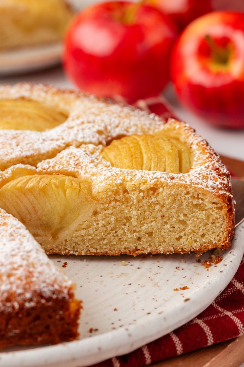 close-up of a slice of German apple cake on a cake plate alongside more cake and fresh apples