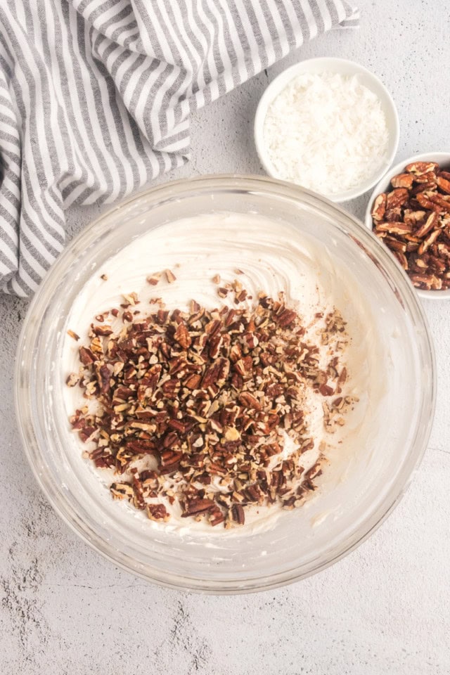 overhead view of chopped toasted pecans added to cream cheese frosting in a glass mixing bowl