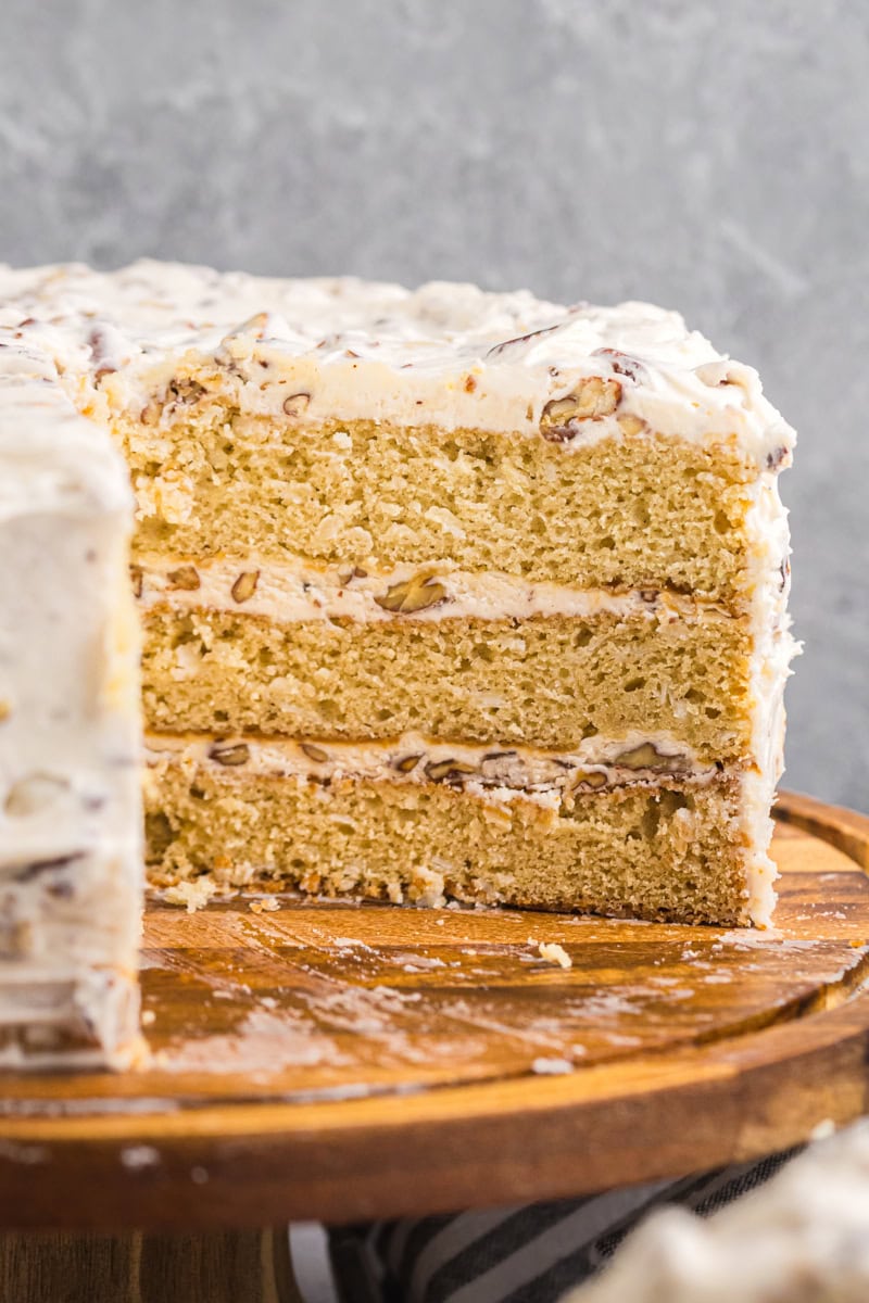 a slice of Italian cream cake on a wooden cake stand, showing three layers of golden cake filled and frosted with cream cheese frosting and pecans