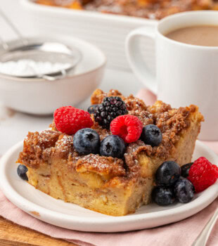 a slice of baked French toast on a white plate with the remaining French toast and a cup of coffee in the background