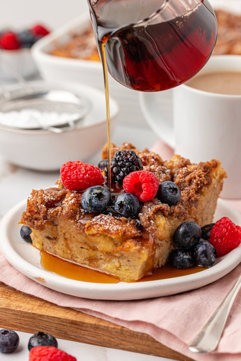 a slice of baked French toast topped with fresh berries with maple syrup being drizzled over it