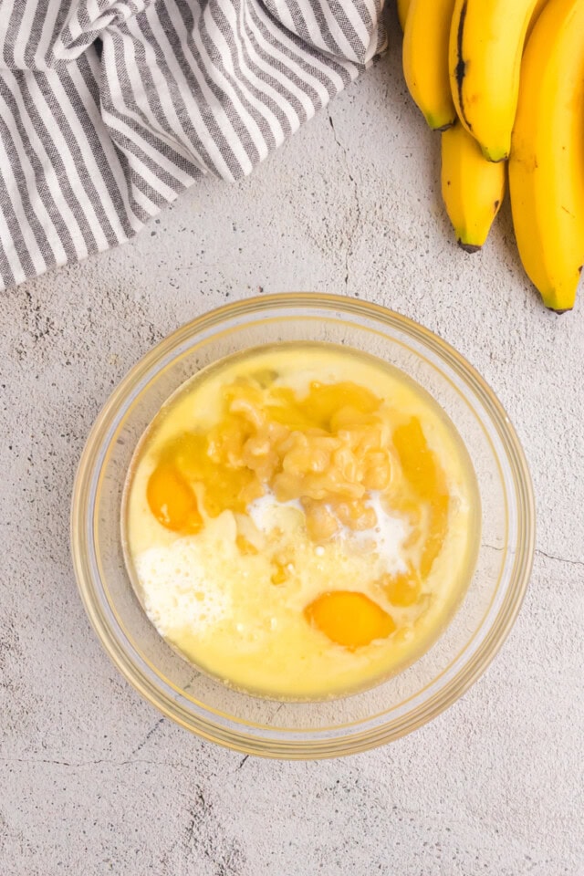 overhead view of melted butter, oil, eggs, vanilla, mashed banana, and milk in a glass mixing bowl