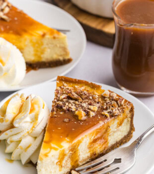 a slice of bourbon caramel swirl cheesecake topped with caramel and nuts, served on a white plate with a dollop of whipped cream; more cheesecake, nuts, and caramel partially visible in the background