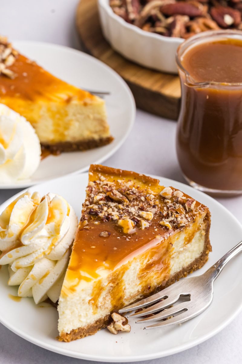 a slice of bourbon caramel swirl cheesecake topped with caramel and nuts, served on a white plate with a dollop of whipped cream; more cheesecake, nuts, and caramel partially visible in the background