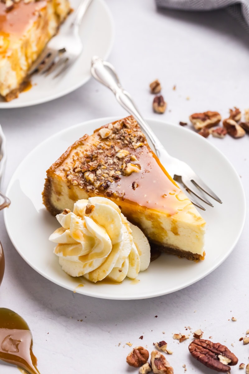 a slice of bourbon caramel swirl cheesecake topped with more caramel and toasted nuts, served on a white plate with a dollop of whipped cream