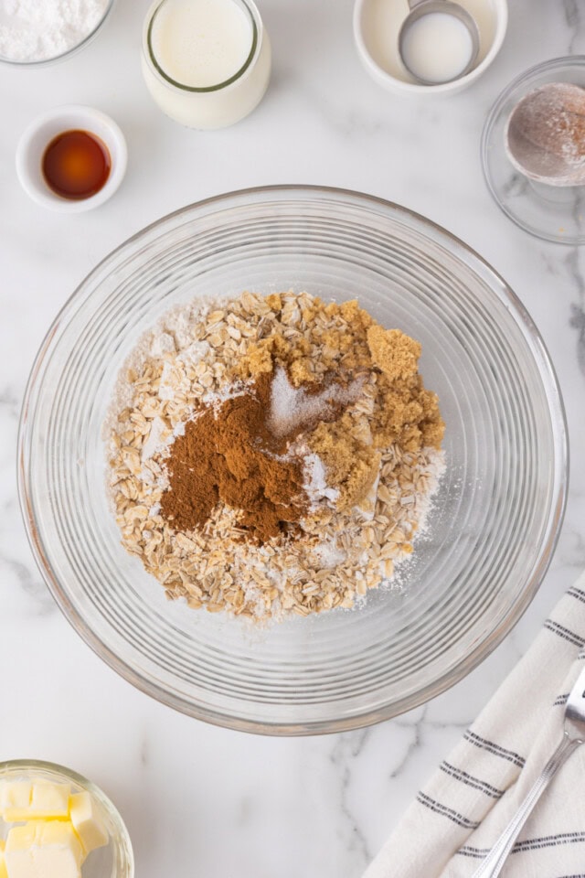 overhead view of flour, brown sugar, oats, baking powder, baking soda, salt, and cinnamon in a glass mixing bowl