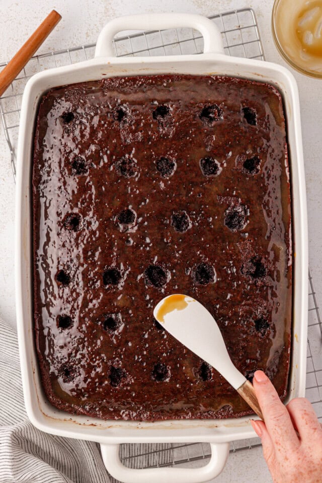 overhead view of caramel being spread over chocolate cake with holes poked into it