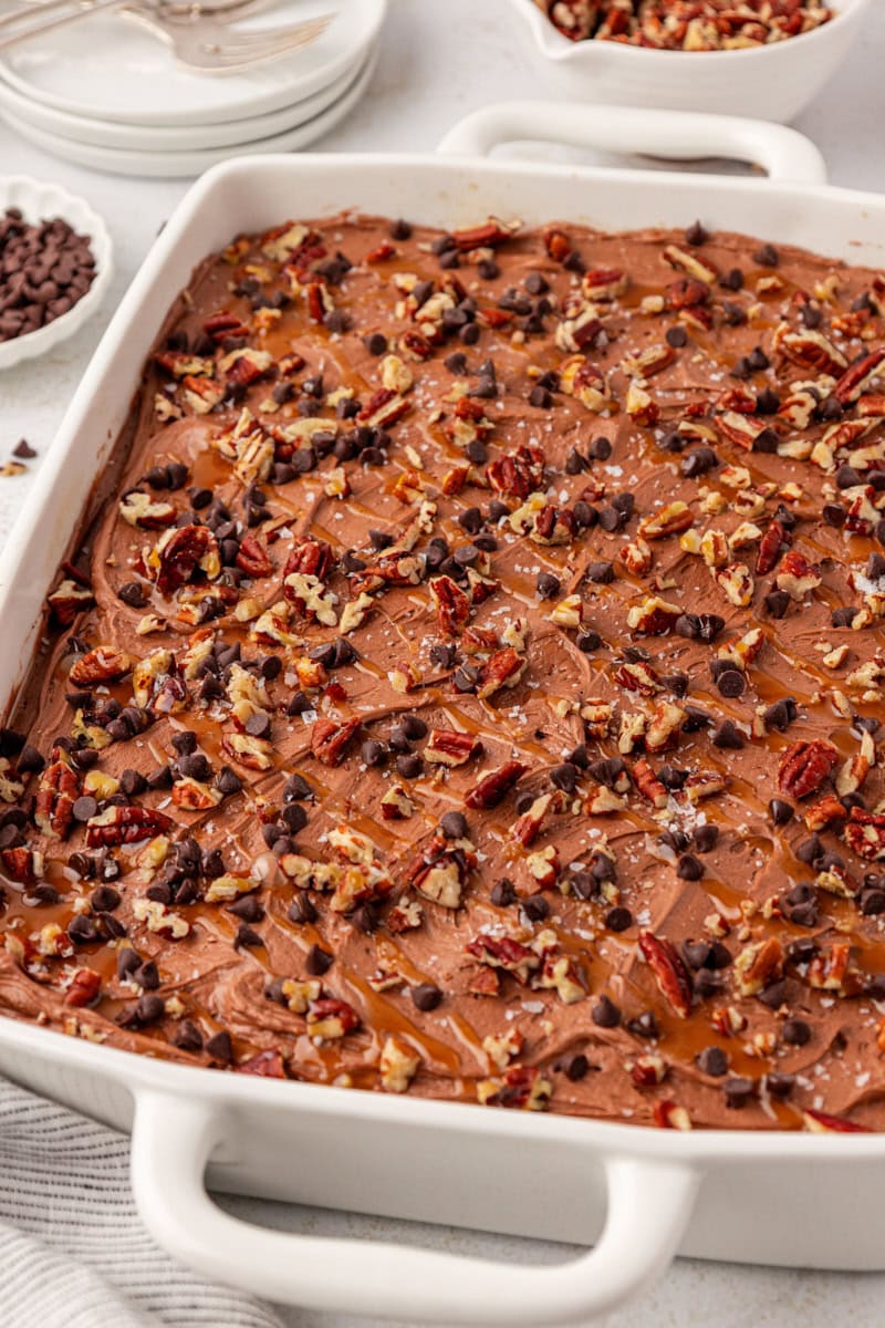 turtle cake topped with caramel, toasted pecans, and mini chocolate chips in a white rectangular baking dish