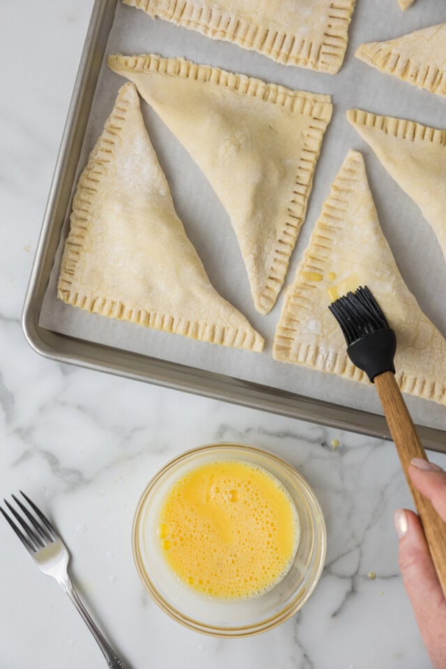 overhead view of egg wash being brushed over apple turnovers on a sheet pan