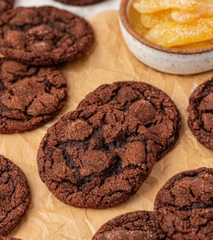 several chocolate ginger cookies scattered on parchment paper with a bowl of candied ginger in the background