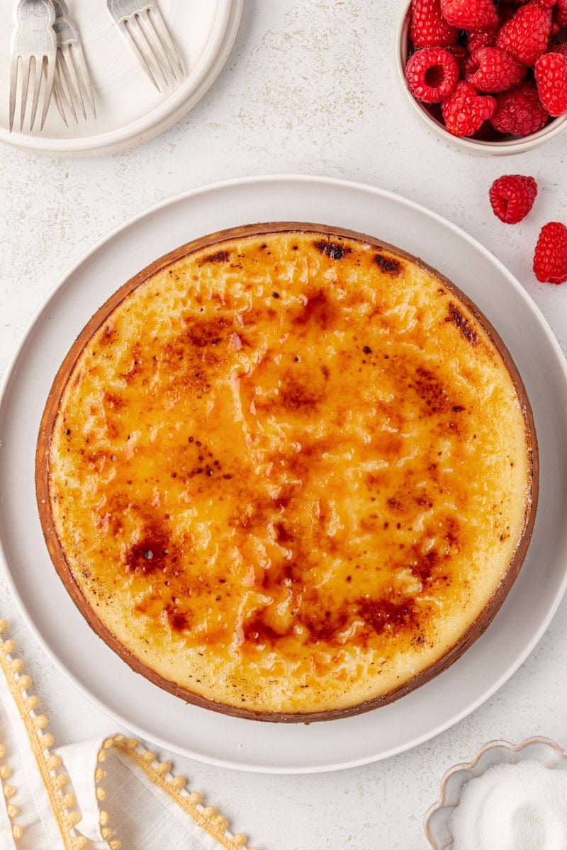 overhead view of crème brûlée cheesecake on a white serving plate, surrounded by plates, forks, and fresh raspberries
