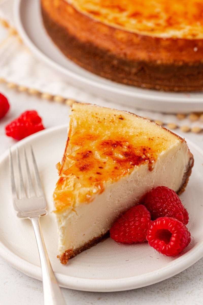 a slice of crème brûlée cheesecake on a white plate with three fresh raspberries and a fork resting on the plate