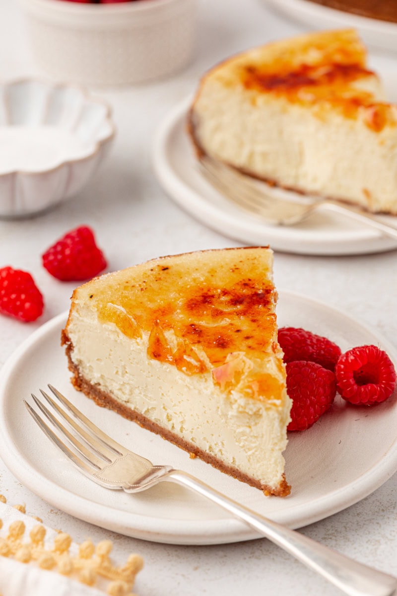 a slice of crème brûlée cheesecake on a white plate with another slice and fresh raspberries in the background