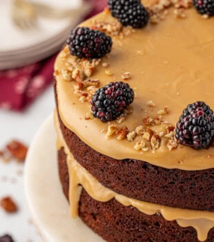 A two-layer Kentucky Jam Cake on a marble cake stand, topped with brown sugar caramel frosting, fresh blackberries, and chopped pecans, with frosting gently dripping down the sides.
