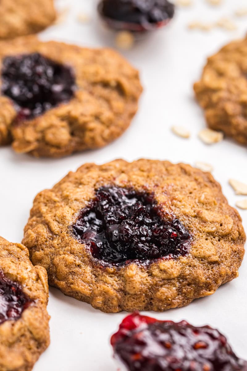 closeup of an oatmeal thumbprint cookie filled with blackberry jam on a white surface with more cookies in the background