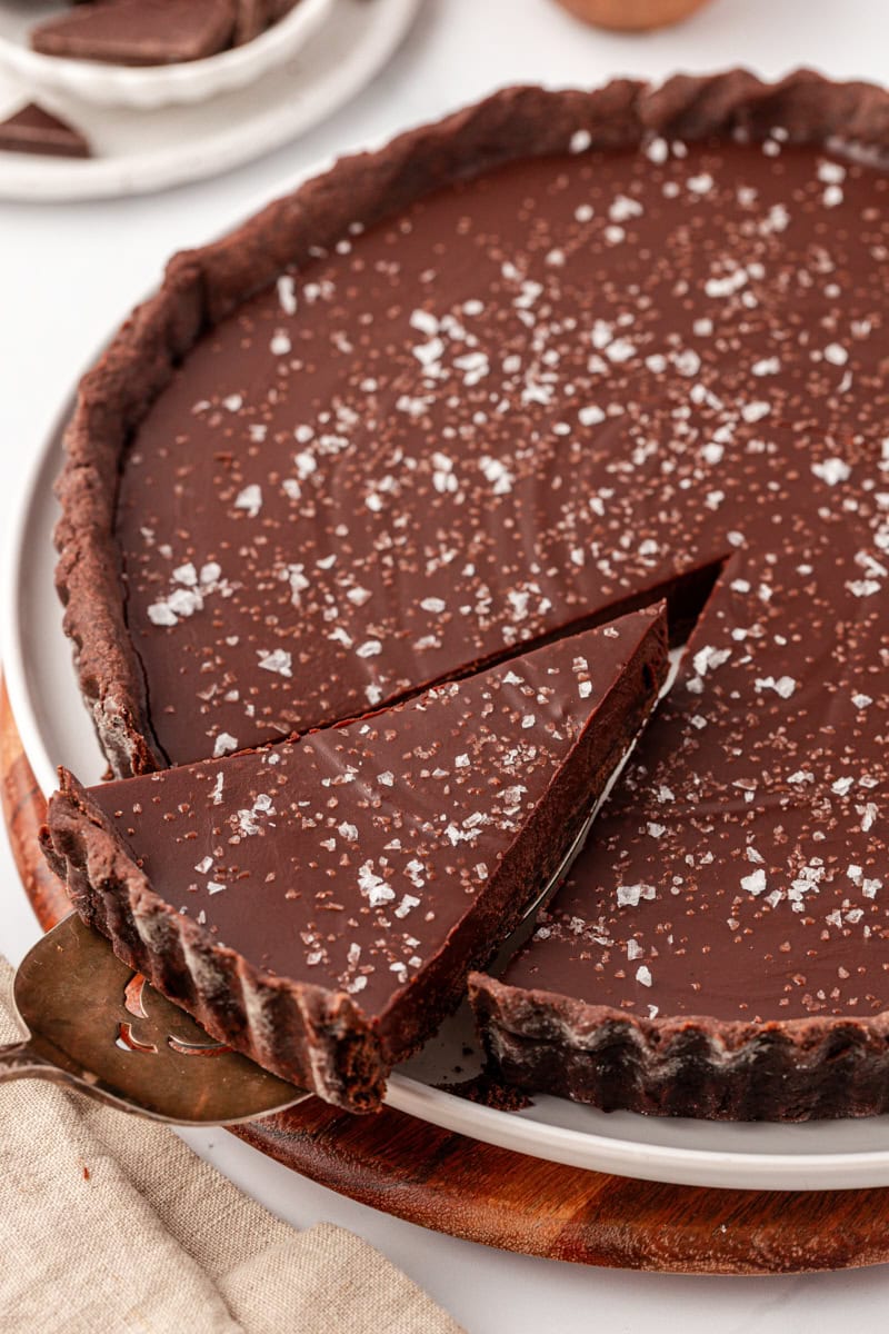 a slice of salted dark chocolate tart on a pie server being lifted away from the remaining tart