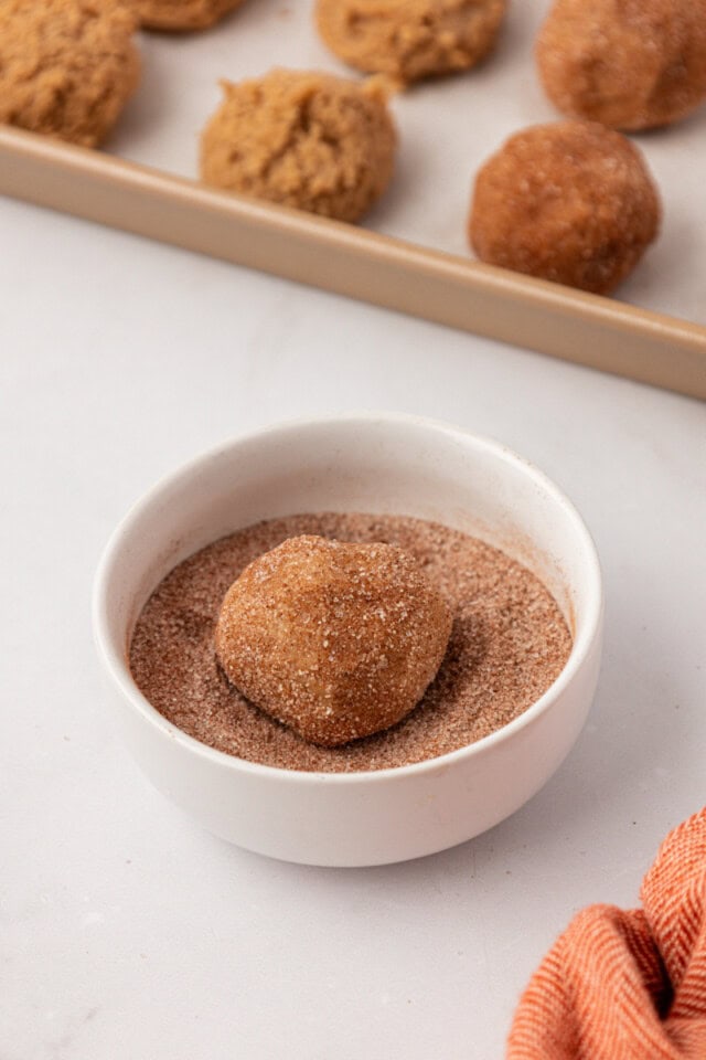 a sugar and spice cookie dough ball being rolled in a bowl of cinnamon-sugar