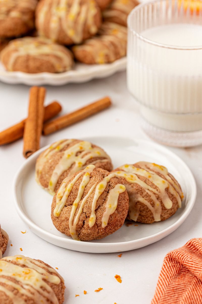 three sugar and spice cookies on a white plate with more cookies and cinnamon sticks in the background