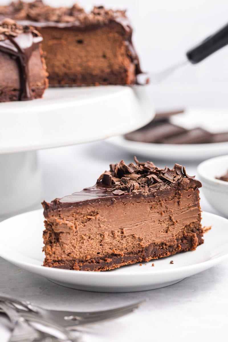 a slice of brownie bottom chocolate cheesecake on a white plate with the remaining cheesecake on a white cake stand in the background