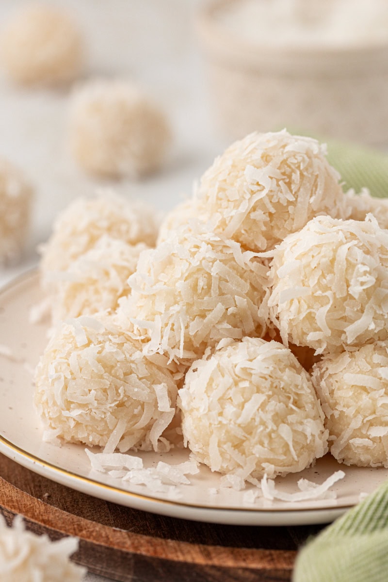 several coconut snowballs piled on a white plate with more snowballs in the background